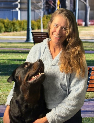 A female professional dog trainer with long wavy hair sits on a wooden bench in a sunny Huntington Beach park, gently holding an attentive Rottweiler during obedience training, showcasing effective positive reinforcement techniques in a calm outdoor setting in Orange County, CA.